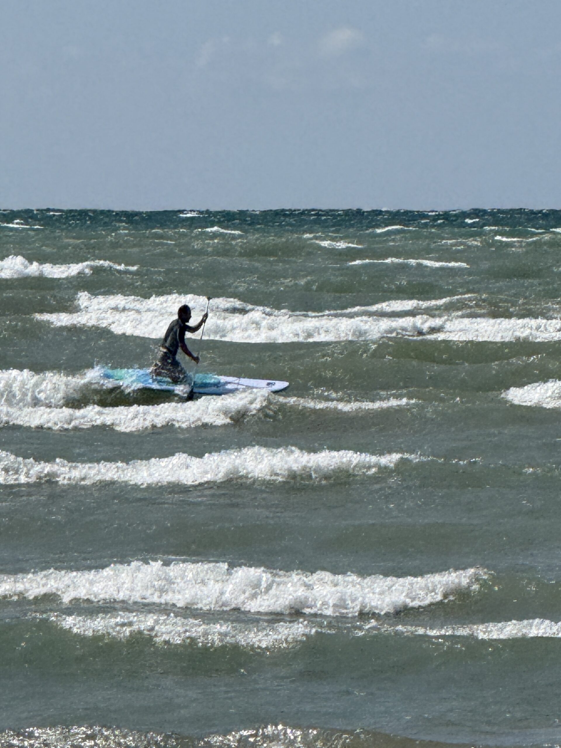 A paddleboarder kneels on a board amid choppy water and breaking waves.